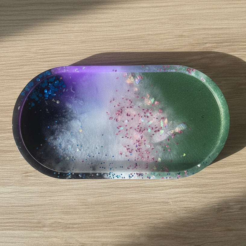 An oval resin tray with a galaxy design, featuring a gradient of black, purple, white, and green with pink glitter, resting on a light wood surface.
