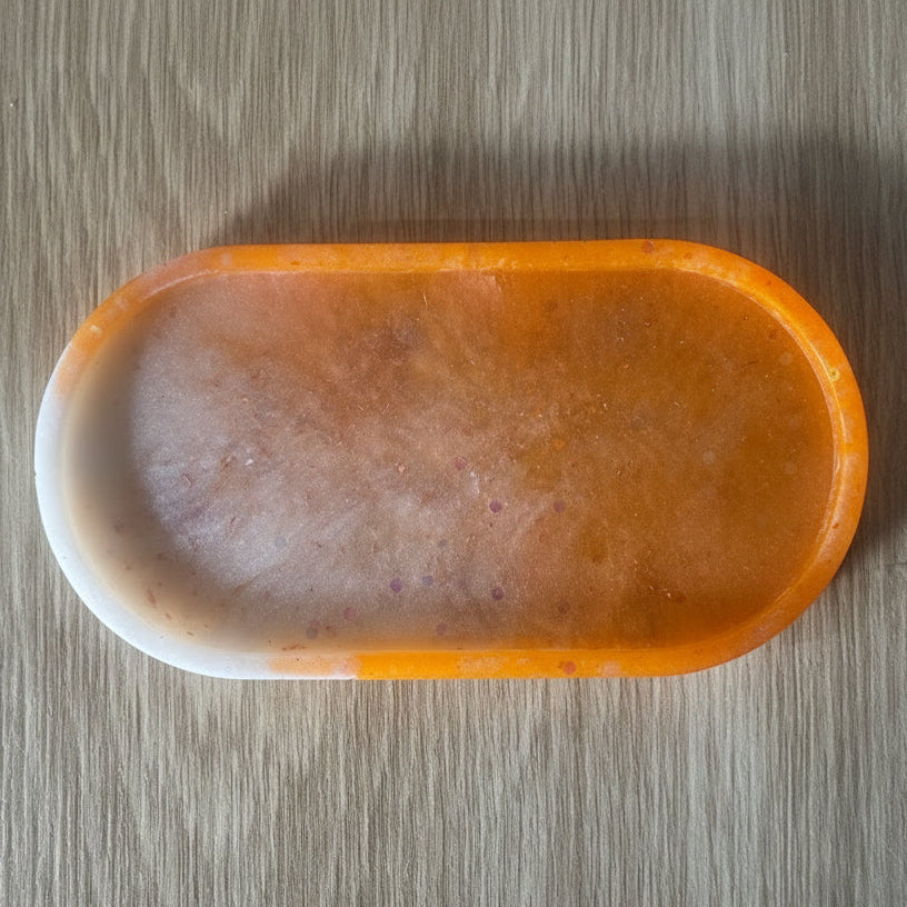 An oval-shaped soap bar with a marbled orange and white color pattern, photographed from directly above on a light wood grain surface.